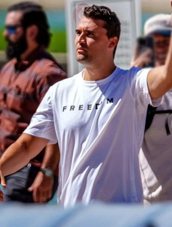 Charlie Kirk wearing a white t-shirt with 'FREEDOM' text, standing outdoors with blurred people in the background.